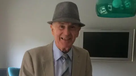 Mackey family An elderly man smiles for a photo. He is wearing a grey hat and a cream jacket and light blue shirt. 