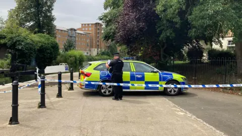 Pete Cooper/BBC A police car is parked across a footpath near a river. A police officer is checking paperwork with his back to the camera. Blue and white police scenes of crime tape has been placed in front of him attached to a metal bollard to the left and iron fencing to the right.
A block of flats is visible in the background on the other side of the river.