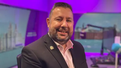 Anthony Burns is sitting in a purple branded BBC Local Radio studio. He has short dark hair, has a short beard and is wearing a shirt and jacket.