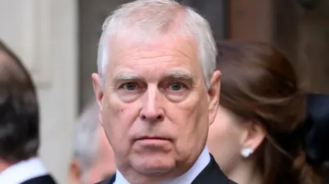 Andrew Mountbatten-Windsor in a black suit, white shirt and black tie looks directly at the camera while standing among other formally dressed people.