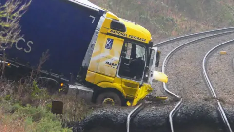 The yellow front cab of a lorry smashed into railway tracks. Two rails are bent and the front tyres and lower part of the cab are lodge in the stones below the track. The rear of the lorry is on the embankment.