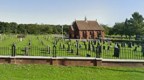 Google Thorpe Road Cemetery on a sunny day. There are lots of graves on green grass and a brick building in the background of the cemetery as well as green trees. There is a brick wall in front and leaves on the grass.
