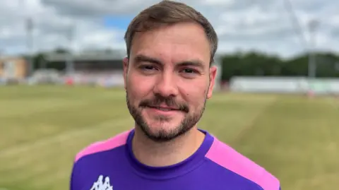30-year-old man with brown hair and facial hair at the Batley Bulldog's rugby league stadium wearing a purple and pink rugby top smiling at the camera.
