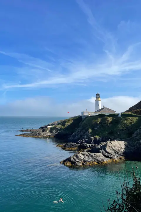 SARAH CALLOW A white lighthouse on a sea cliff on a sunny day. There is someone swimming in the sea below, which is greenish in colour and the sky above is blue with a streak of cloud across it.