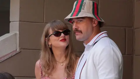 Taylor Swift, wearing a red gingham top, looking up at Travis Kelce, wearing a striped Gucci bucket hat