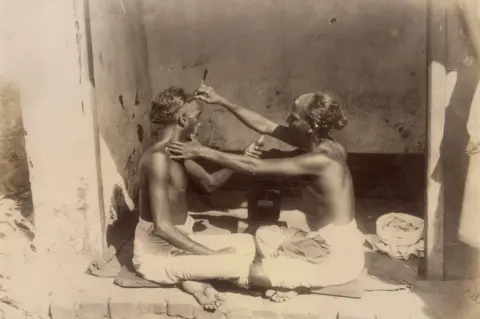 DAG A street barber trims a man’s hair in a small vacant space in India. Both wear traditional dress - a sarong-type dress. A turban lies near a man. The photograph was taken by an unidentified photographer.
