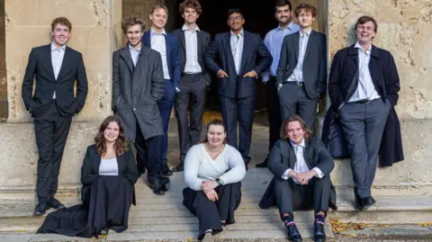 Music in Action Eleven members of choir Liliam stand together wearing dark suits and smart clothes, outside a large, historic building.