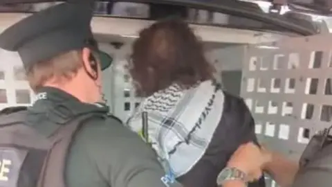 Other Screengrab of dark haired woman being loaded into the back of a police van. She is wearing a black and white scarf, probably a keffiyeh, and has a dark coat on. Two police officers, one more visible, are helping her into the van.