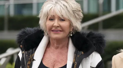 PA Media A middle aged woman with short blonde hair, red lipstick and wearing a black and white coat with a black fur-lined hood, smiles into the camera.