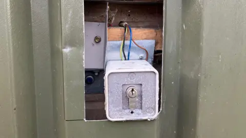 Guy Campbell/BBC A rectangular hole has been cut in a green metal casing exposing a lock attached to yellow, black, blue and brown wires.