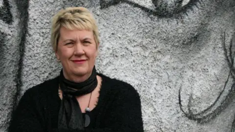 Vici Wreford-Sinnott looking into the camera and smiling. She has short, blonde hair and is wearing pink lipstick, a black scarf and black jumper. She is in front of a grey mural which looks to show a face.