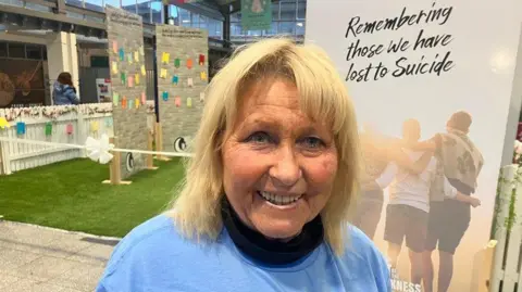 Eileen Morgan, a woman with blond hair, wearing a blue shirt. She is standing in front of a pull-up banner with a photo of three people embracing and black text reading "remembering those we have lost to suicide".