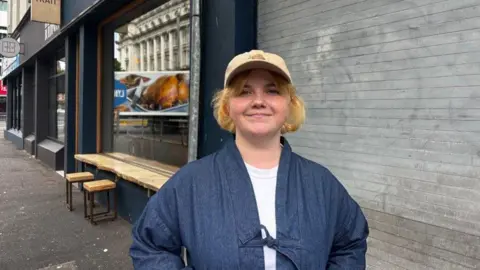 A young girl on the street, smiling. Wearing a white top, denim jacket and yellow cap.