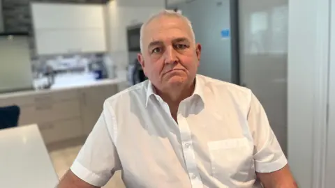 A man with short grey hair wears a short sleeve white shirt. Behind him, out of focus, is a kitchen with white floors and white counter tops.