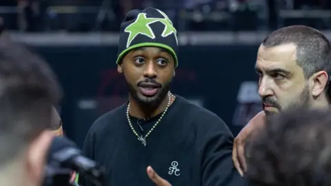 Baller League Sharky is in a huddle with players on the pitch. He wears a black sweatshirt, green and black beanie hat and a light-coloured necklace