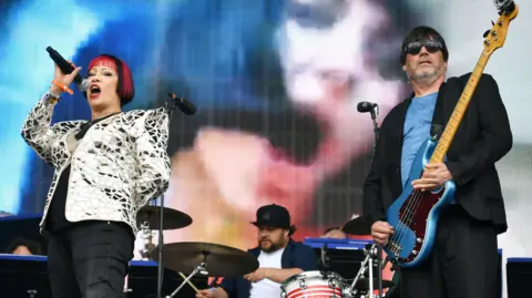 Jim Dyson/Getty Images A woman singing onstage alongside a man who is playing a blue bass guitar. 