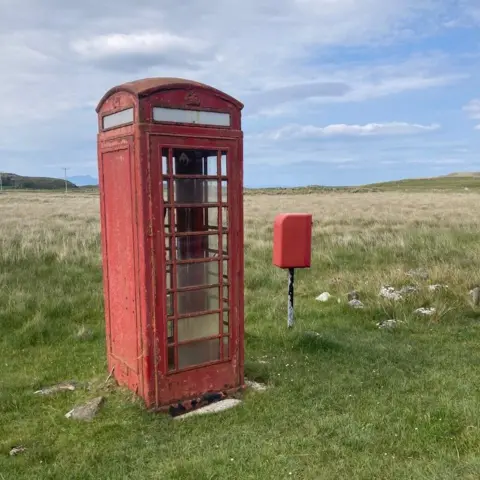 KATH MANNERS A red phonebox in a grassy field. It has been damaged and has bits of paint coming off it. A small red postbox facing away from the camera is to its right. They are both on a grass patch with longer grass behind it.