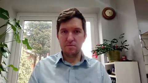A man with short brown hair sitting in an office with pot plants in the background and trees visible through the window behind him. He is wearing a blue shirt. 