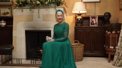 Getty Images Helen Mirren sits in front of the mantelpiece, wearing a long emerald green dress and a matching hairband.