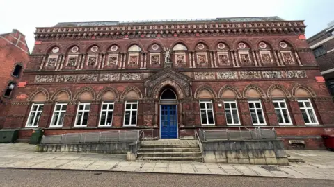 The Wedgwood Institute, a large red-brick building with a series of arched windows on its ground floor and carved figures within archways on the second floor. There are blue double doors in the centre of the building with steps and a ramp leading up to them.