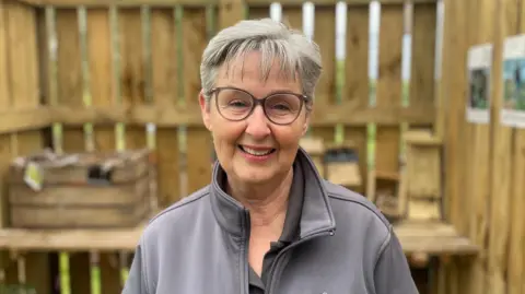 A woman has short grey hair and wears clear glasses with grey frames. She smiles at the camera as she stands inside a wooden hut wearing a grey zip-up jacket. 