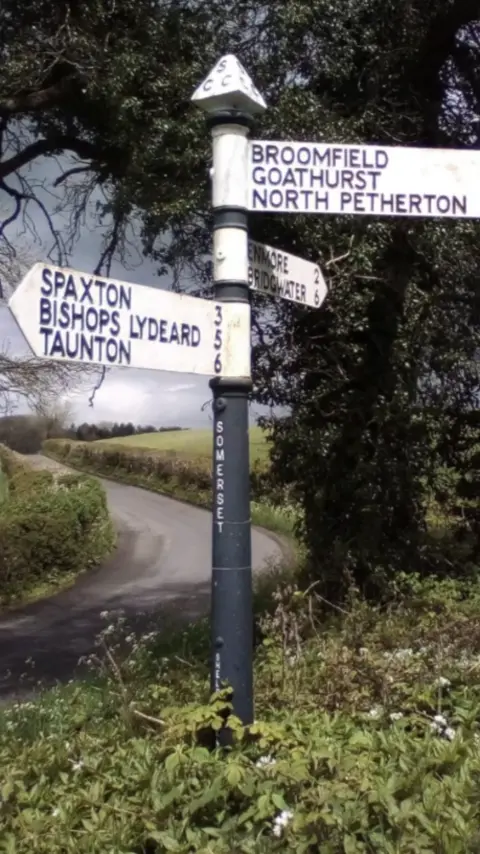 A white signpost pointing in three directions, positioned next to a curved country road.