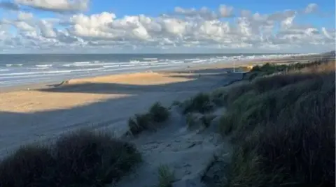 The photograph is looking out over the Belgian coastline with sand dunes in the foreground, then a sandy beach, before the Channel coast comes into view.