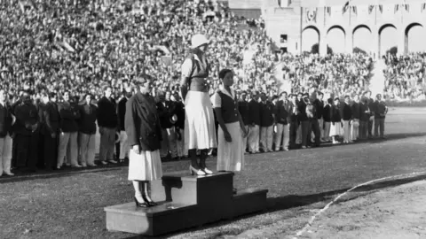 O holandês Willie Den Oden, as medalhistas de ouro Helen Madison dos EUA e Eleanor Saville dos EUA estão juntos no pódio da vitória nos 100 metros livres nas Olimpíadas de 1932