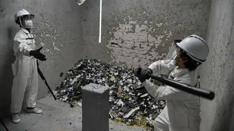 Getty Images Picture of two people in a rage room smashing items with baseball bats. They are wearing hard hats, visors and white overalls.
