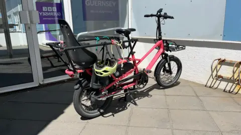 A red cargo e-bike is parked on a brown paved surface opposite a wall coloured in white, gray and blue. There is a rusty bike rack attached to the same wall. The bike has bungee straps attached to its frame and a yellow cycle helmet is attached to the frame and hangs upside down over the lower right hand side. To the left of the bike there is a glass double door with purple coloured BBC Guernsey signs attached. 