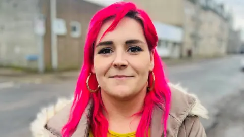 Skye Ryan, woman with bright pink hair, smiling at camera, wearing a beige jacket with fur-trimmed hood, and buildings in the background.