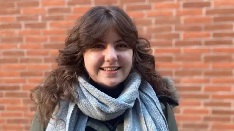 A woman with long brown hair in a blue scarf smiles.