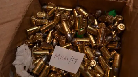 ERSOU A cardboard box filled with gold-coloured bullet casings. A white card is lying in the box with "MBM/17" written on it. What appears to be a piece of tissue paper is also in the box.