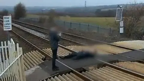Network Rail A person wearing black is lying down on the railway track. Their face is blurred. Another person in black is standing and taking a photo on a phone.