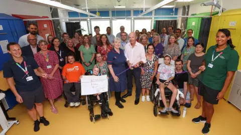 Southampton Children’s Hospital A large group photo shows dozens of people gathered in a hospital room to mark the launch of Robbie's Rehab. In the centre, Robbie's dad Mark stands holding a pair of scissors against a purple ribbon. He is bald and wears a pale pink shirt with dark blue jeans and black leather shoes. Mark stands next to a hospital sister, dressed in a dark blue dress uniform. There are two children either side of them sat in wheelchairs. Everyone is smiling at the camera and one child holds up a sign which reads 'Robbie's Rehab'.