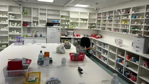Laura Fowler The photo shows dozens and dozens of boxes of medication left on shelves inside the pharmacy after Jhoots were asked to leave. More medication, note books and baskets full of other items can also be seen on a large white table in the middle of the room.