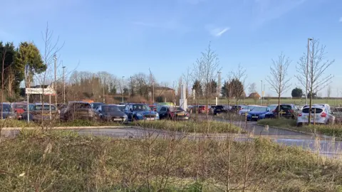 The station car park with rows of cars parked under a blue sky and fields and grass around it. One car is parked on the grass.