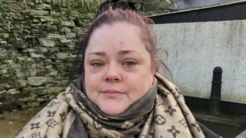 Geraldine Kelly has pink hair, which is tied back. She is wearing a large silk scarf that is brown and grey. She is standing near a stone wall. 