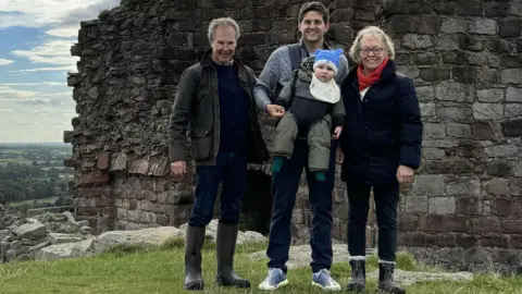 Family photo A family group standing outdoors in front of the remains of an old stone wall, part of an historic ruin or castle. The people are dressed in outdoor clothing, including jackets and boots, and the man in the centre is carrying a small child. Behind the group there is a scenic view of green fields stretching into the distance under a partly cloudy sky. 