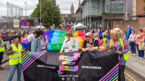 Omagh Pride A group of people carry an Omagh Pride banner. They are part of a parade with bystanders watching. Most people are in brightly coloured clothes.