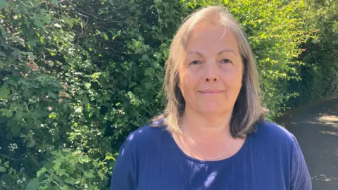 Councillor Sarah Murray has blonde hair and is wearing a blue T-shirt and a necklace. She is standing outside with shrubs behind her.