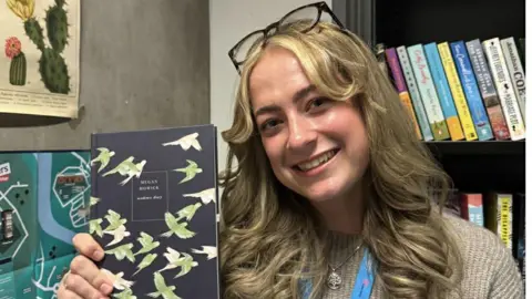 Supplied Megan Howick is smiling as she holds a book. She has long wavy blonde hair and blue eyes. She is wearing a beige jumper and a pair of black-rimmed glasses over the the top of her head. There is a shelf of books behind her. 