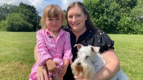 Caitlyn in a pink and white striped dress is sat cuddling a woman with long hair who is wearing a black top with coloured hearts on it. The woman has her other hand around a white dog. Grass and trees can be seen behind them.