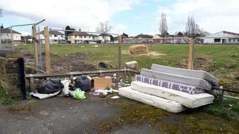 Five mattresses and bin bags dumped on a patch of land with hay and grass behind a metal fence