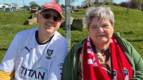 An older man and woman sit side-by-side in front of a grassy area. They are both wearing York City FC football shirts. The man wear a white shirt, a red cap and sunglasses. The woman has short hair, and wears a brown shirt, green hoodie and red scarf.