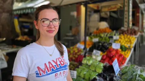 Paul Moseley/BBC Billie Waring is wearing a white t-shirt with 'Maybe Baby' on the front. She has large framed glasses and we can see a selection of vegetables behind her.