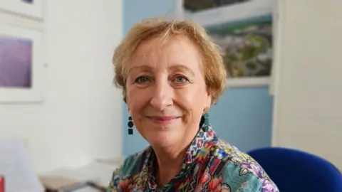 Lesley Dwyer is smiling at the camera and is wearing a colourful patterned top. She has short, light auburn hair and is wearing dangly blue earrings