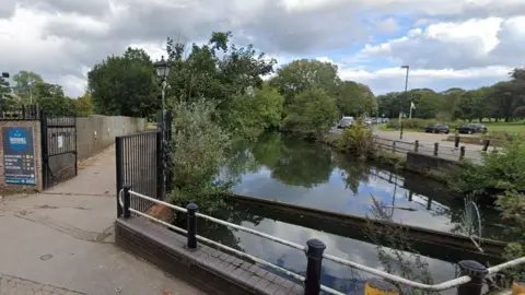 River in centre with road and path either side - the water is still and the river bank edged with trees.