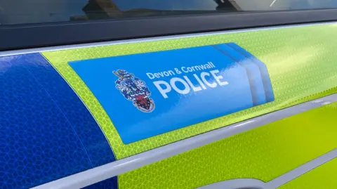 The side of a yellow and blue police vehicle. Above the door handle is a blue section with the words 'Devon and Cornwall Police' with a badge to its left. 