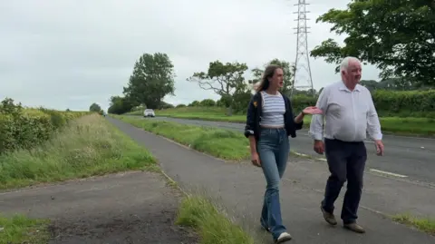 Holly Fox and Peter Gaston walking along a section of footpath next to a main road. She has long dark hair and is wearing a navy cardigan and jeans, while he has short white hair and is wearing a white shirt and navy trousers. A car can be seen in the background. The road and path have grass verges on either side.